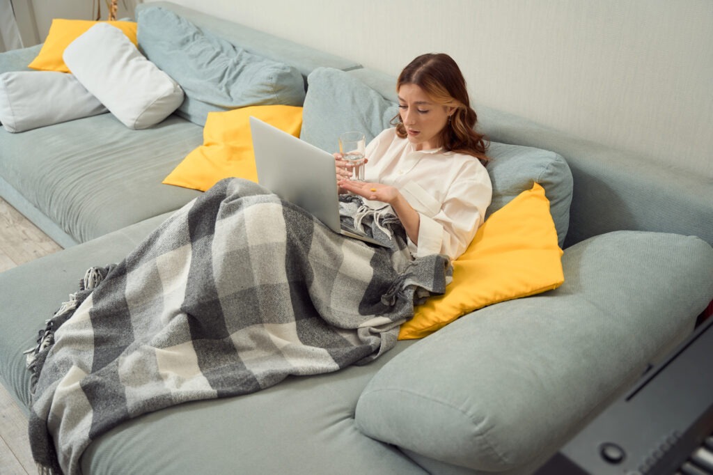 Caucasian woman laying on a pale blue couch under a plaid blanket looking at her laptop.