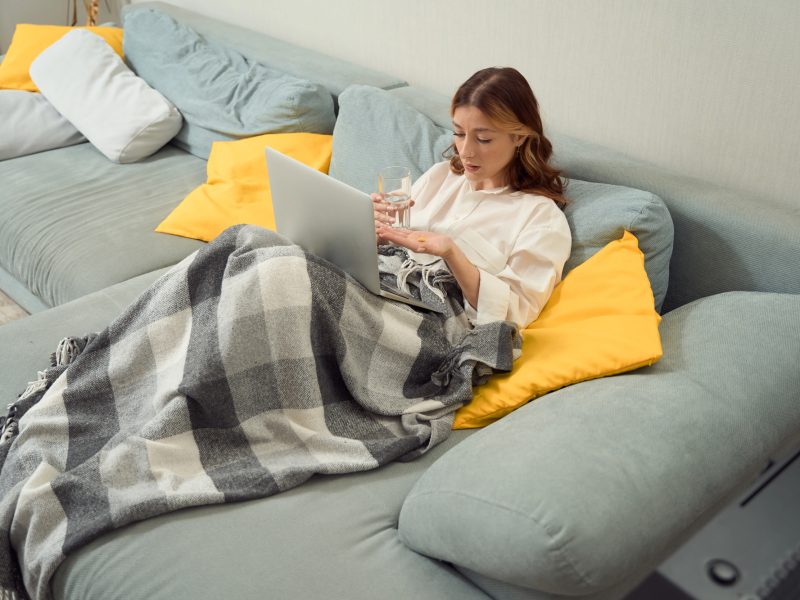 Caucasian woman laying on a pale blue couch under a plaid blanket looking at her laptop.